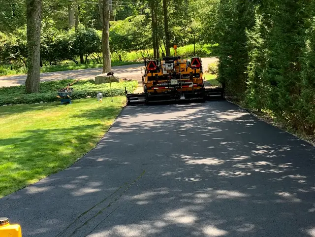 Asphalt paving machine laying a new driveway surface along a wooded lot